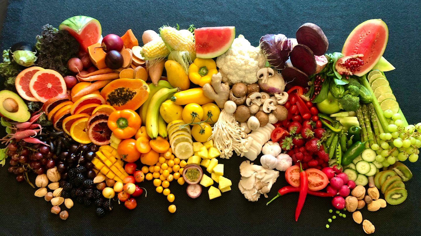 Assortimento di frutta fresca, verdura e noci disposte a arcobaleno su una superficie scura.
